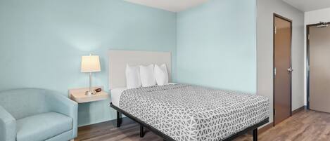 Habitación de hotel con una cama, una silla, una mesita de noche con una lámpara y una puerta.