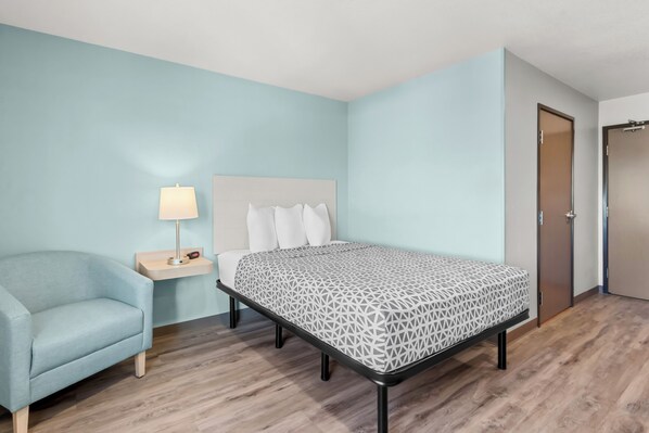 A hotel room with a bed, a chair, a nightstand with a lamp, and a door.