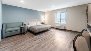 A modern hotel room with a bed, a sofa, a small table, a lamp, a window, and a built-in shelf. - Woodspring Suites Wilmington Riverfront (Wilmington)