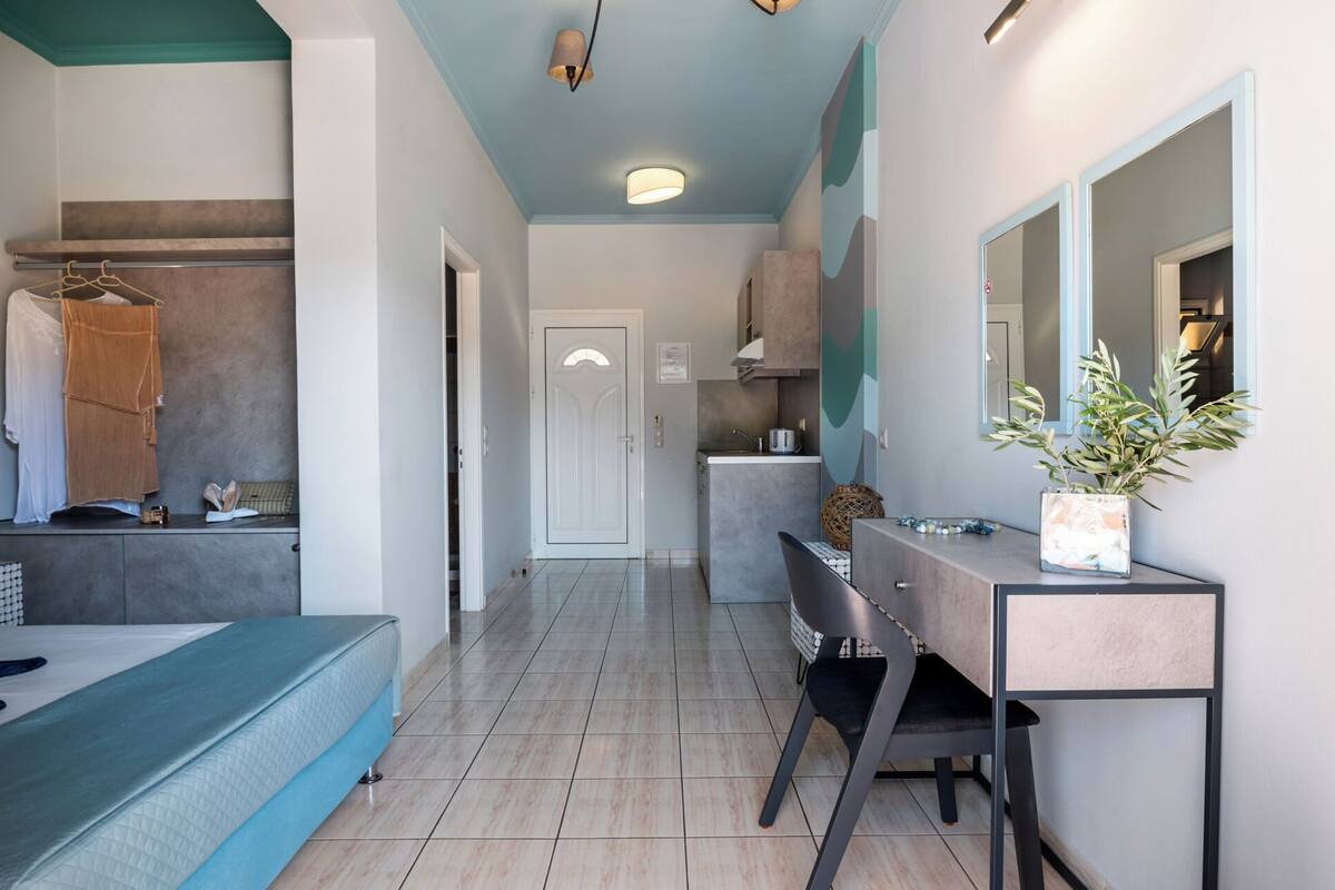 A modern hotel room with a bed, a desk with a chair, a mirror, and a small kitchenette.