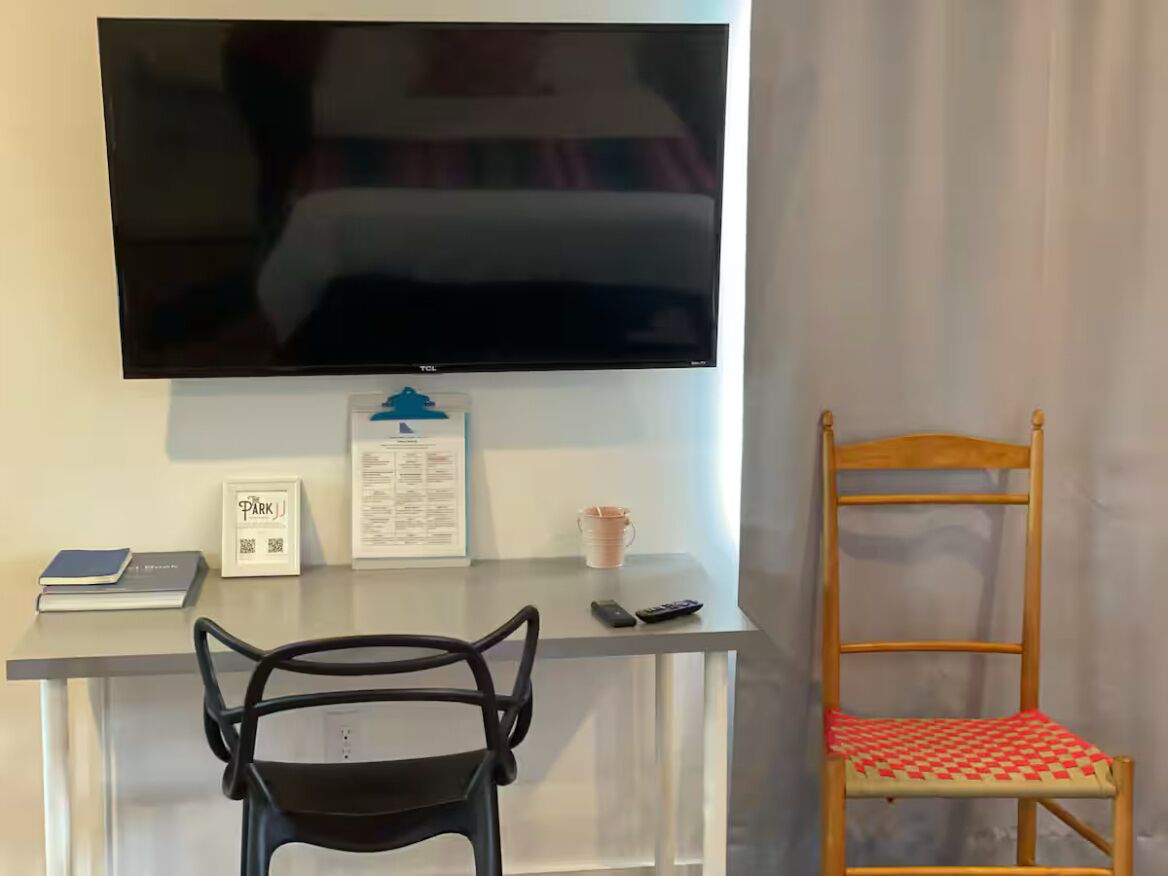 A room with a TV mounted on the wall, a desk with a chair, a wooden chair with a checkered cushion, and a framed sign on the wall.