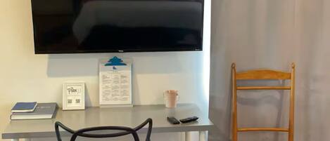 A room with a TV mounted on the wall, a desk with a chair, a wooden chair with a checkered cushion, and a framed sign on the wall.