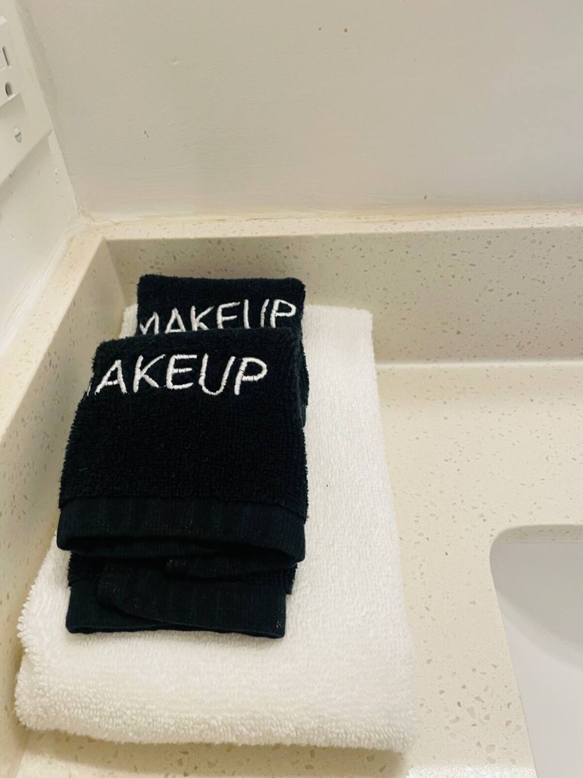 Een stapel zwarte handdoeken met de tekst 'MAKEUP AKEUP' erin geborduurd, geplaatst op een witte handdoek in een badkameringe.