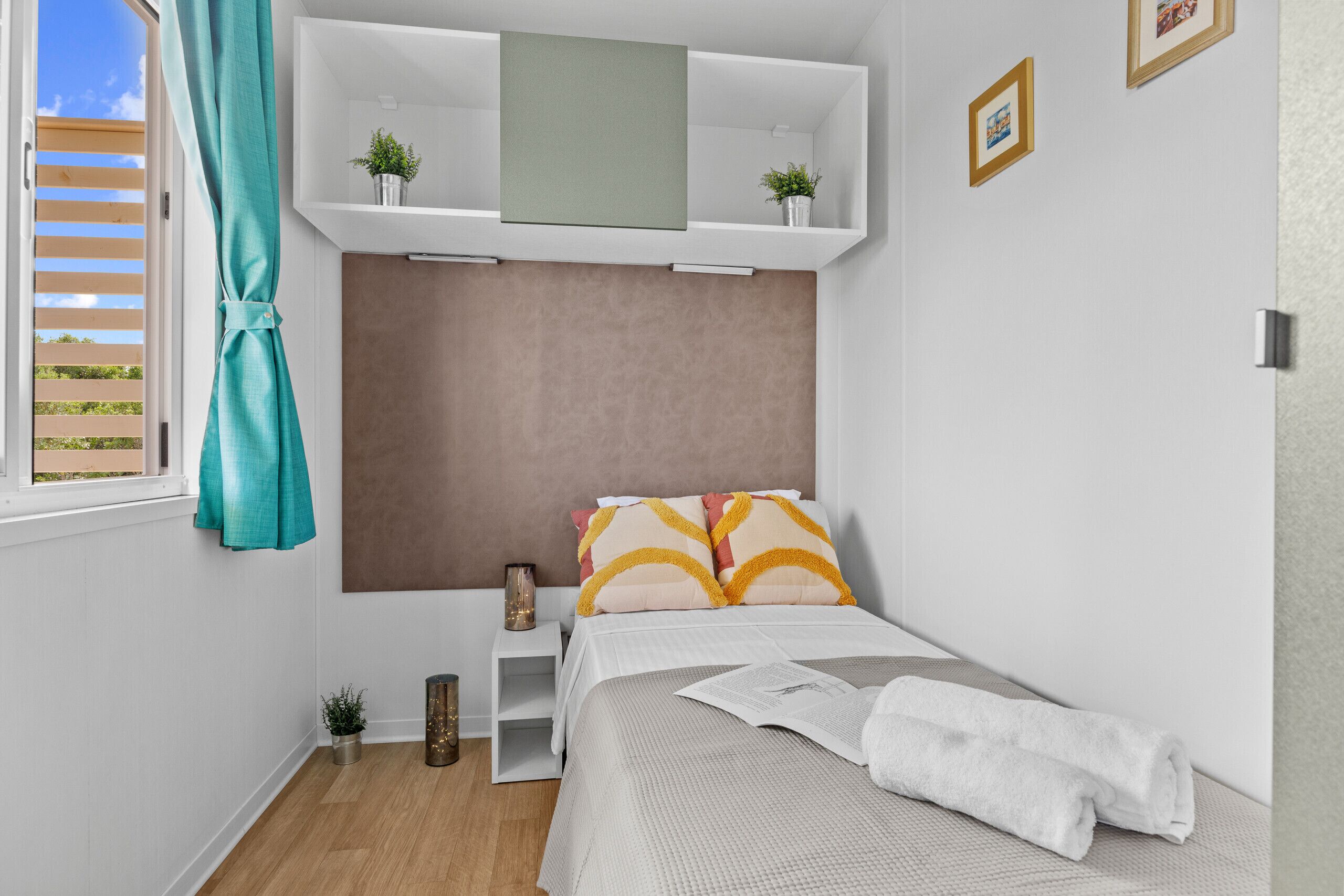 A bedroom with a bed, a window with curtains, a shelf with plants, and a bedside table with a lamp.