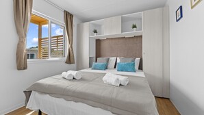 Une chambre avec un grand lit, deux tables de chevet, une fenêtre avec des rideaux et une étagère intégrée où sont disposées des plantes.