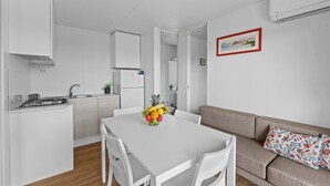 Une cuisine moderne avec des meubles blancs, une table à manger avec des chaises, un panier de fruits et un canapé agrémenté d’un coussin à motifs.