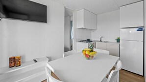 Une cuisine moderne avec des meubles blancs, une table à manger avec des chaises, un panier de fruits et un téléviseur à écran plat fixé au mur.