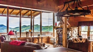 Interior de una cabaña de madera con un ventanal grande, una chimenea y un comedor.