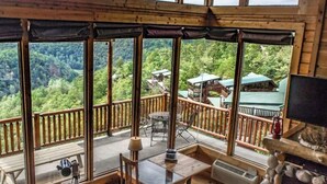 Una terraza de madera con una mesa y sillas, ventanas amplias con vistas a un bosque y cabañas, y una chimenea de piedra.