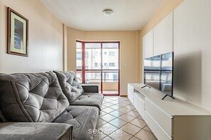 A living room with a grey sofa, a TV on a white cabinet, and a balcony with a view of buildings. - Apt Foot in the Sand sea View in Meia Praia Evb308 (Itapema)