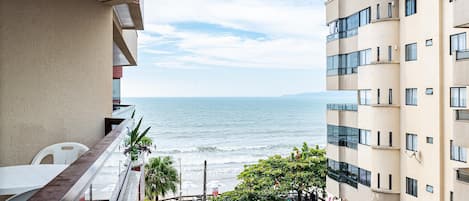 Un balcón con vista a una playa, donde se ve el mar, un árbol y un edificio de varios pisos.
