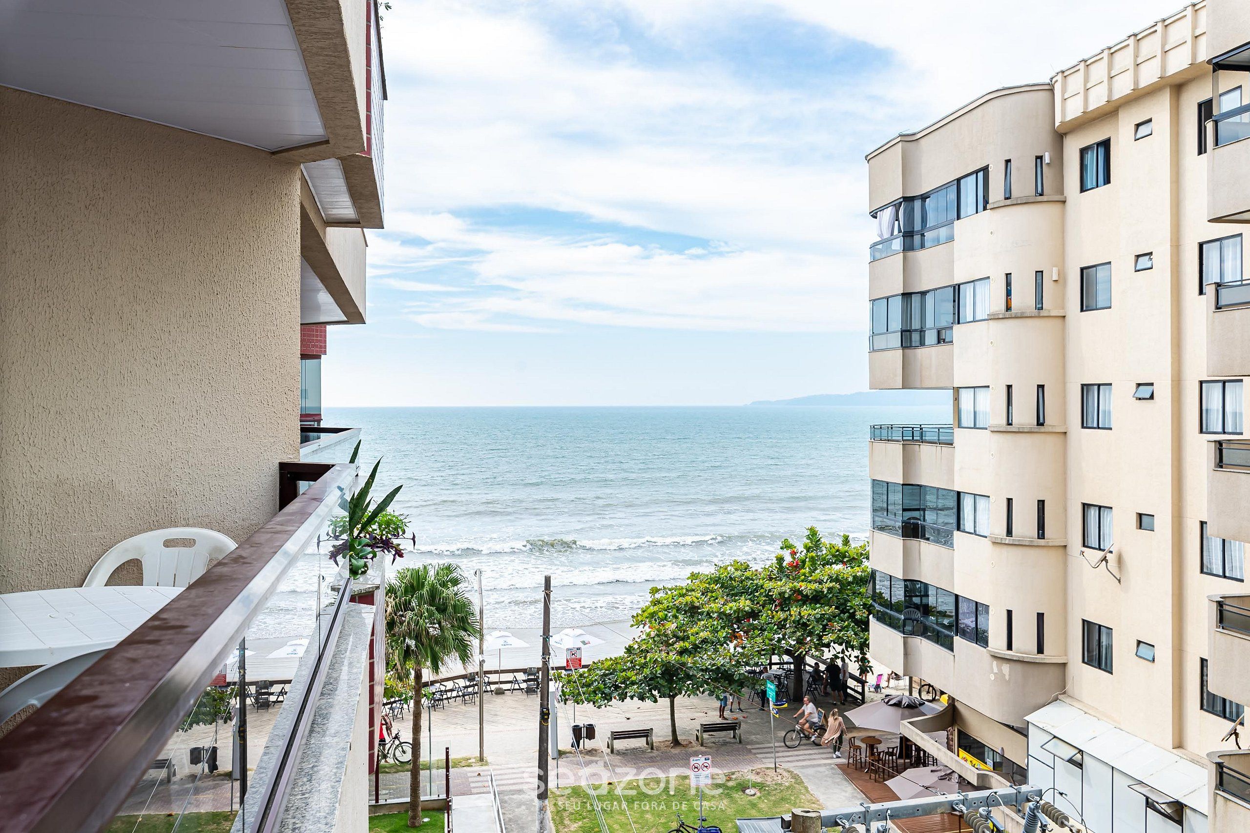 Un balcón con vista a una playa, donde se ve el mar, un árbol y un edificio de varios pisos.
