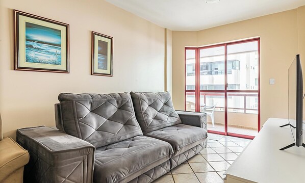 Un salon avec un canapé en cuir gris, deux tableaux encadrés au mur, un téléviseur posé sur un meuble et un balcon avec une rambarde rouge.