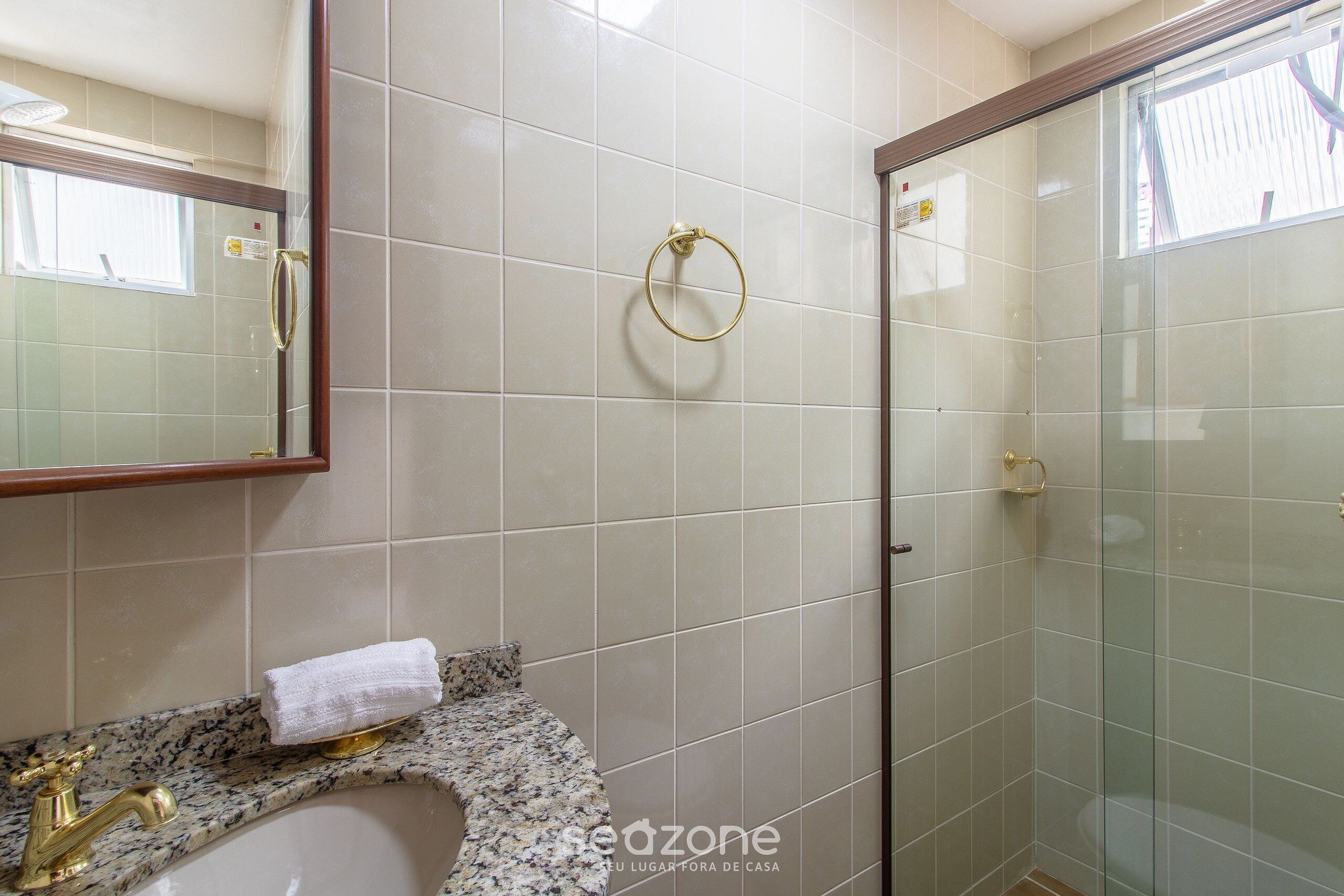 A bathroom with a granite countertop, a gold faucet, a towel, a glass shower enclosure, and a mirror with a wooden frame.