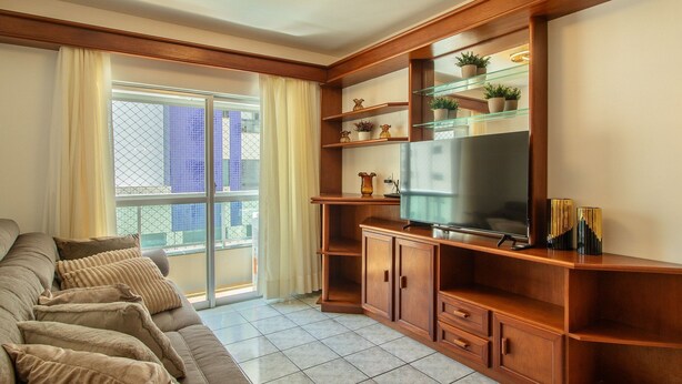 A living room with a sofa, TV, wooden shelves, and a balcony with a view of buildings. - 3BR Apt w/ Balcony 100m From Beach Evz0601 (Balneário Camboriú)