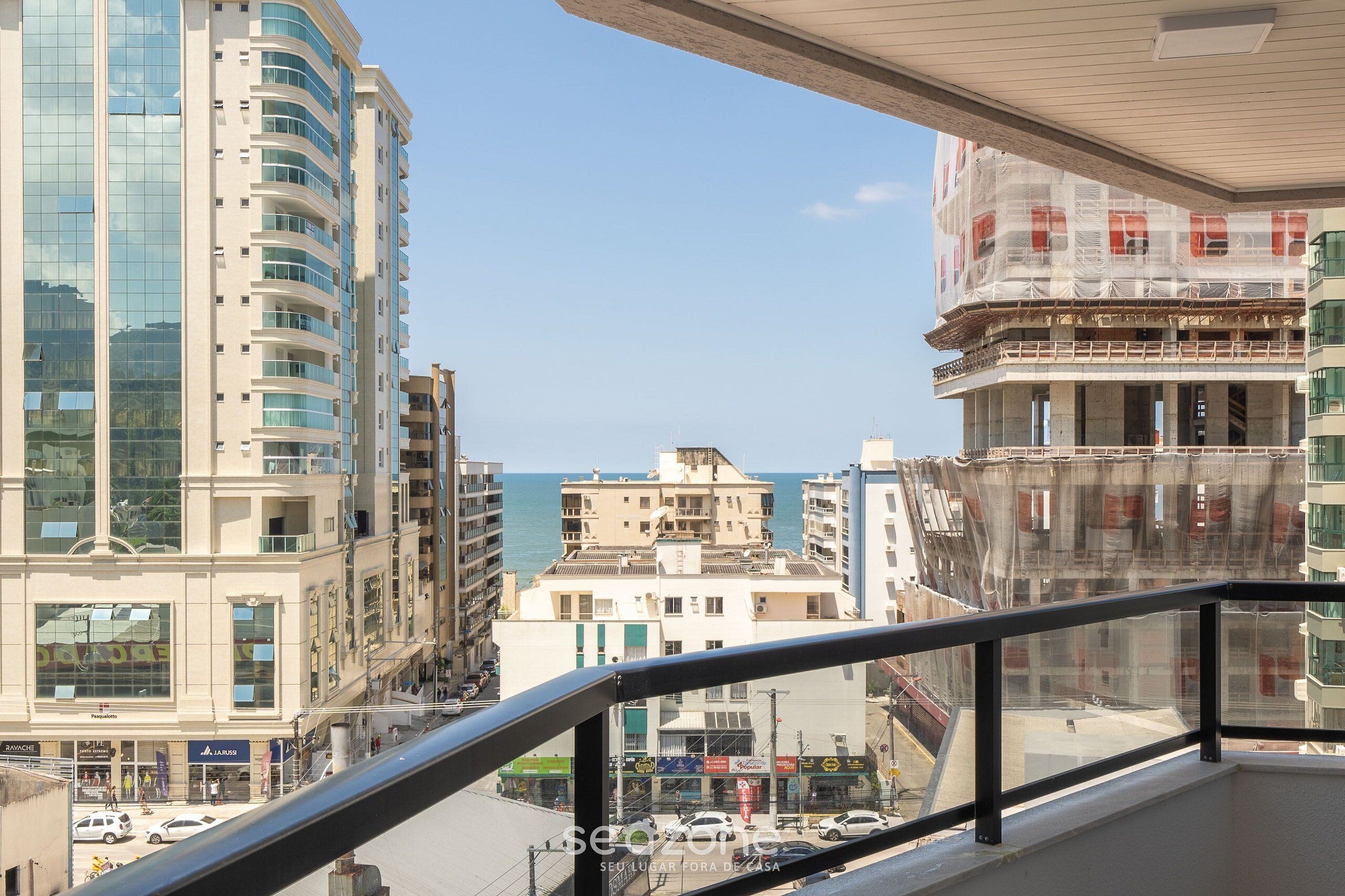 Ein Hochhaus mit Glasfassade, ein Balkon mit Metallgeländer und Blick auf das Meer sowie weitere Gebäude im Hintergrund.