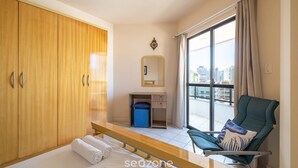 A hotel room with a bed, a chair, a wardrobe, a desk, a mirror, and a window with a city view. - Apt With sea View and Barbecue in Itapema Gbl601 (Itapema)