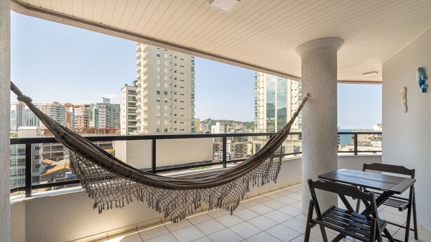 A balcony with a hammock, a table, and chairs, overlooking a cityscape with tall buildings. - Apt With sea View and Barbecue in Itapema Gbl601 (Itapema)