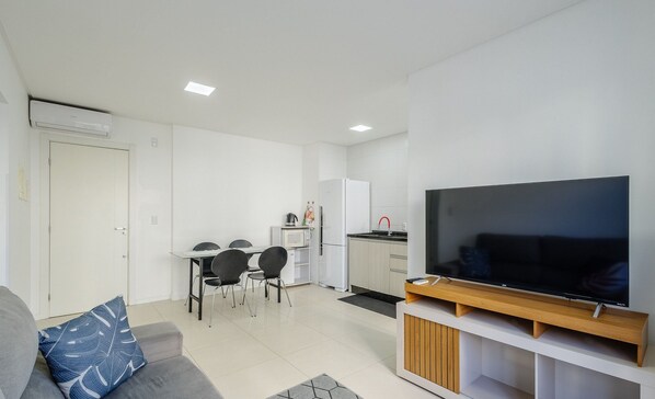 A modern living room with a flat-screen TV, a grey sofa with a blue pillow, a dining area with chairs and a table, and a kitchen with a microwave and refrigerator.