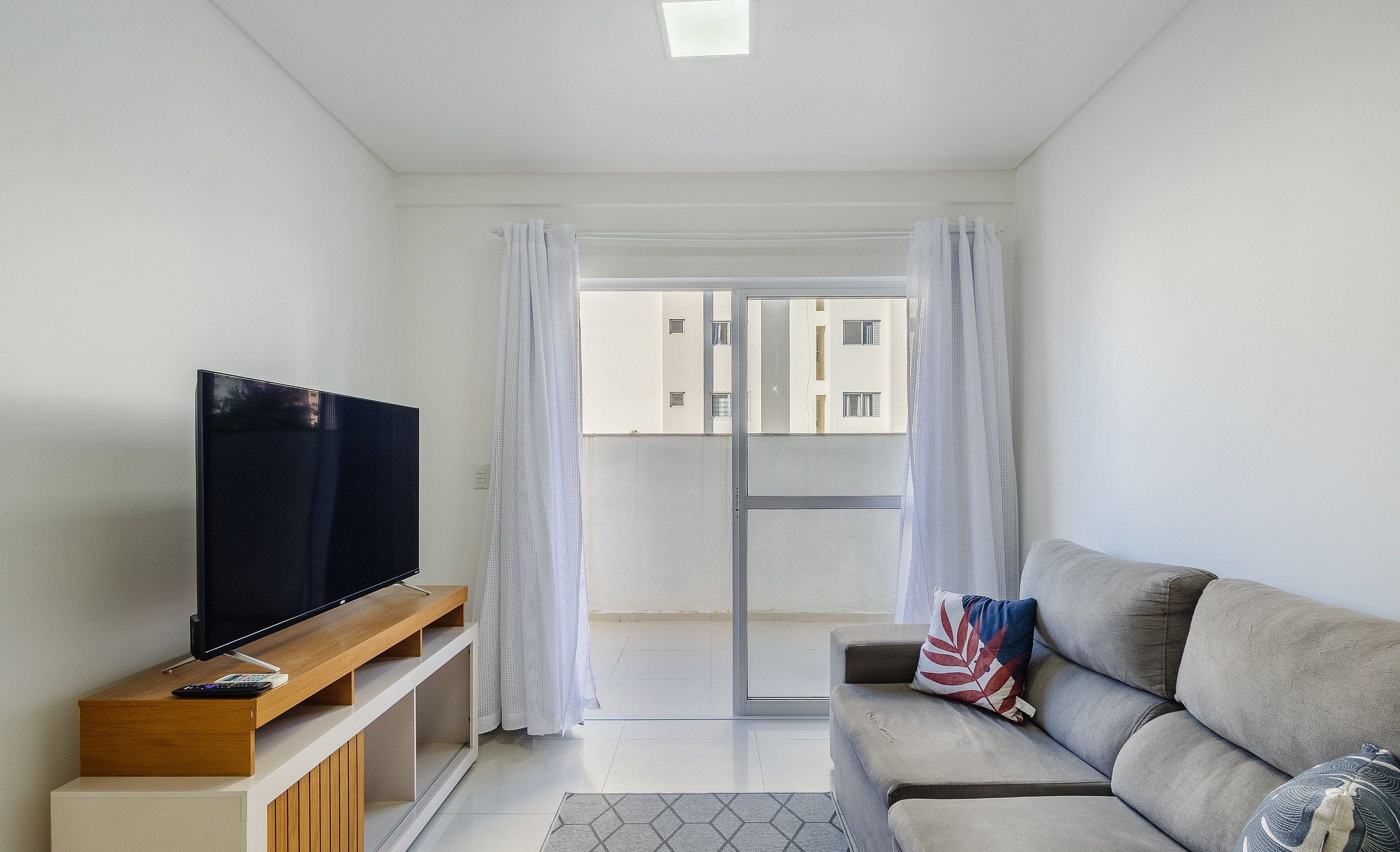 Uma sala de estar moderna com televisão de tela plana em suporte de madeira, sofá cinza com almofadas decorativas e porta de vidro deslizante para uma varanda externa.