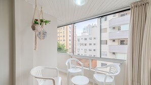Un balcón con sillas de plástico blanco y una mesa redonda pequeña, un colgador de planta de macramé, y vista a edificios de varios pisos.