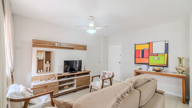 Una sala de estar con televisor, mueble de entretenimiento de madera, ventilador de techo, un cuadro abstracto colorido y un sofá color beige.