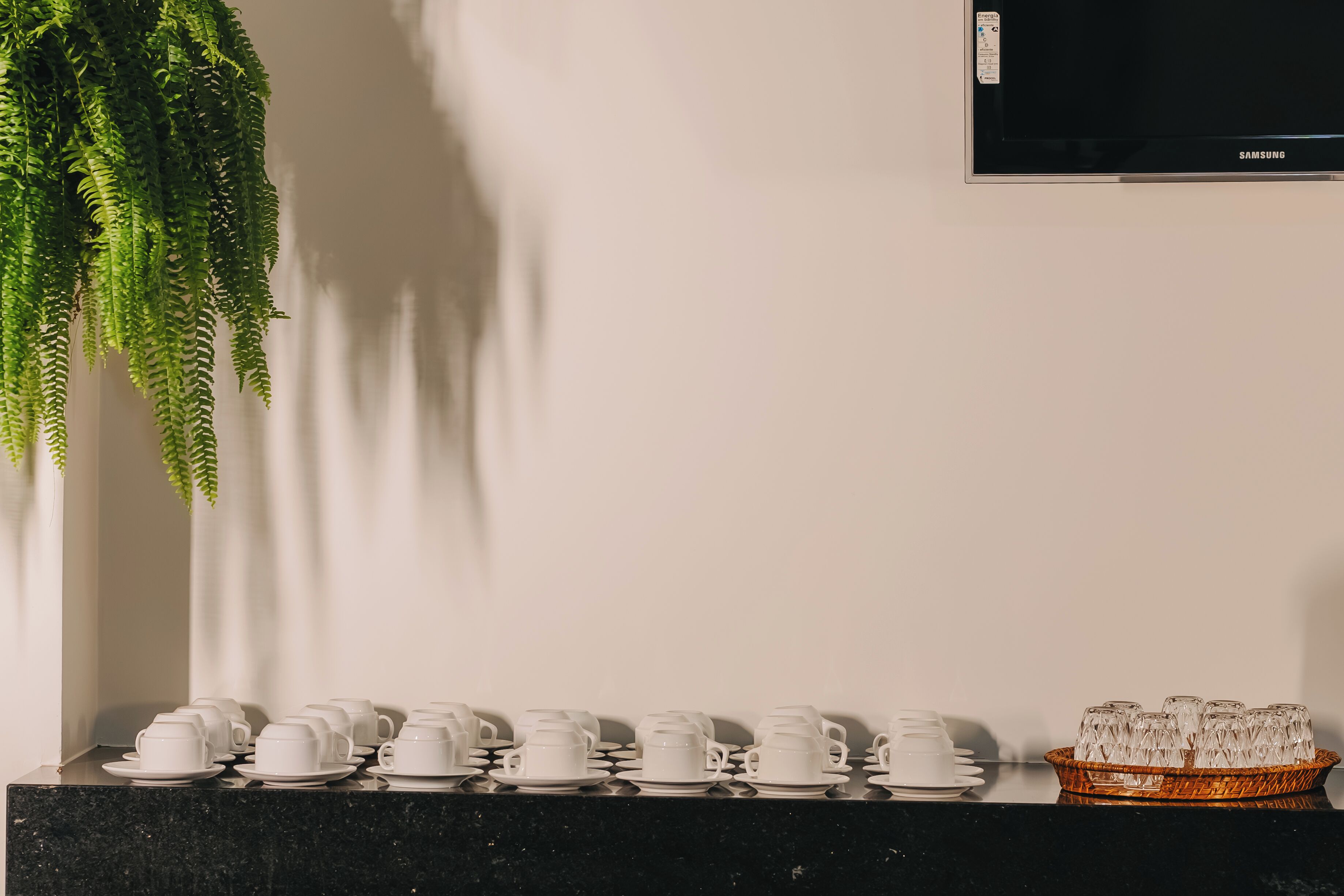 A room with a black marble countertop, white ceramic cups and saucers, and a wicker basket with clear glasses.