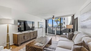 A modern living room with a large flat-screen TV, wooden cabinets, a sofa, and a balcony with a view of the city. - Three-bedroom, two-bath condo at Anglers Cove. Partial view of water and pool. (Marco Island)