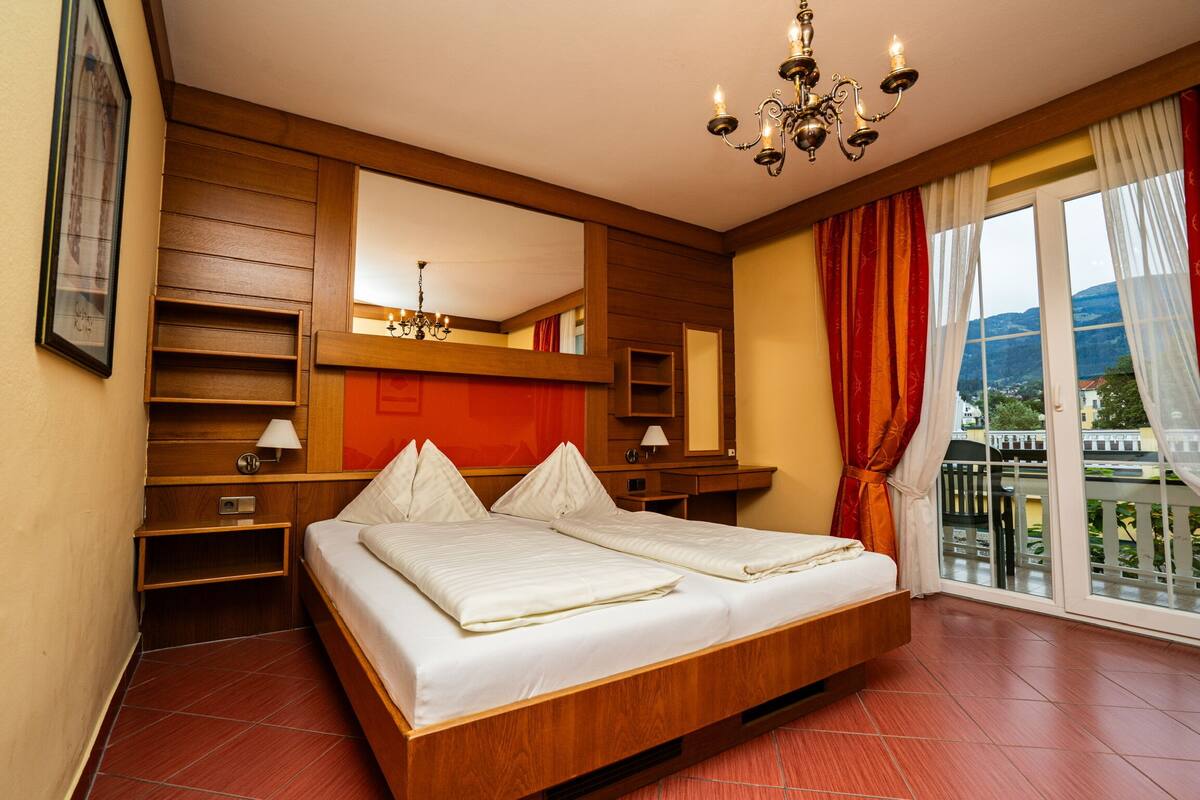 A hotel room with a large bed, wooden paneling, a chandelier, a large window with curtains, and a view of the outside.