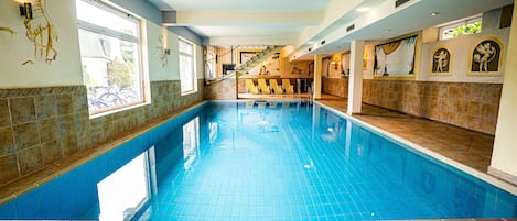 An indoor swimming pool with tiled walls and a tiled floor, a staircase leading to a balcony, and a bar area with yellow chairs.