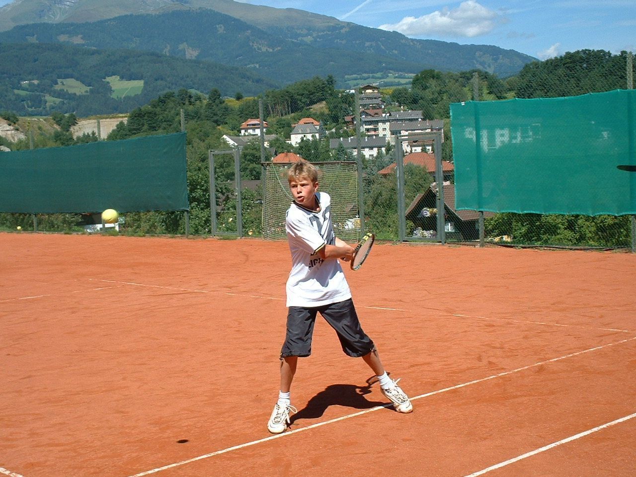 Una cancha de tenis con superficie de arcilla, rodeada de vallas verdes y con casas y montañas al fondo.