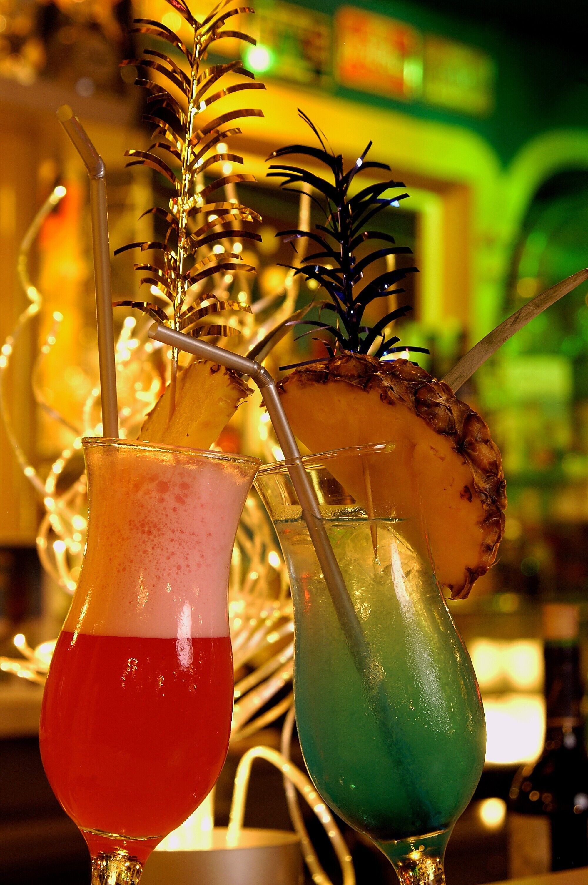 Two cocktails with straws and a pineapple garnish, in a bar setting with a blurred background.