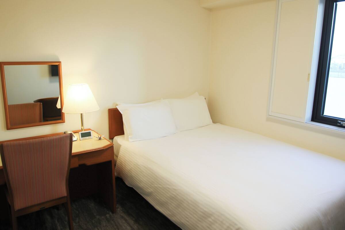 A hotel room with a bed, a bedside table with a lamp, a chair, a mirror, and a window.