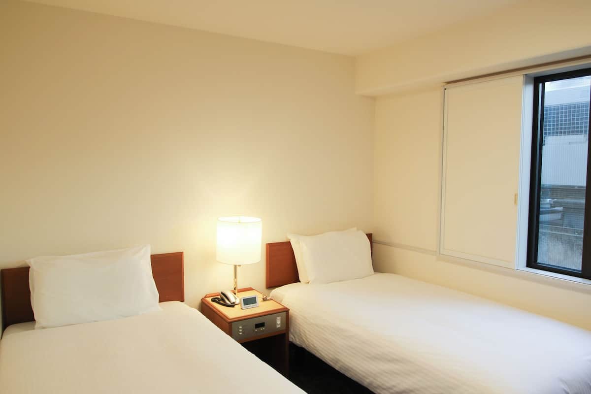 A hotel room with two single beds, a bedside table with a lamp, a phone, and a window with a view of the city.