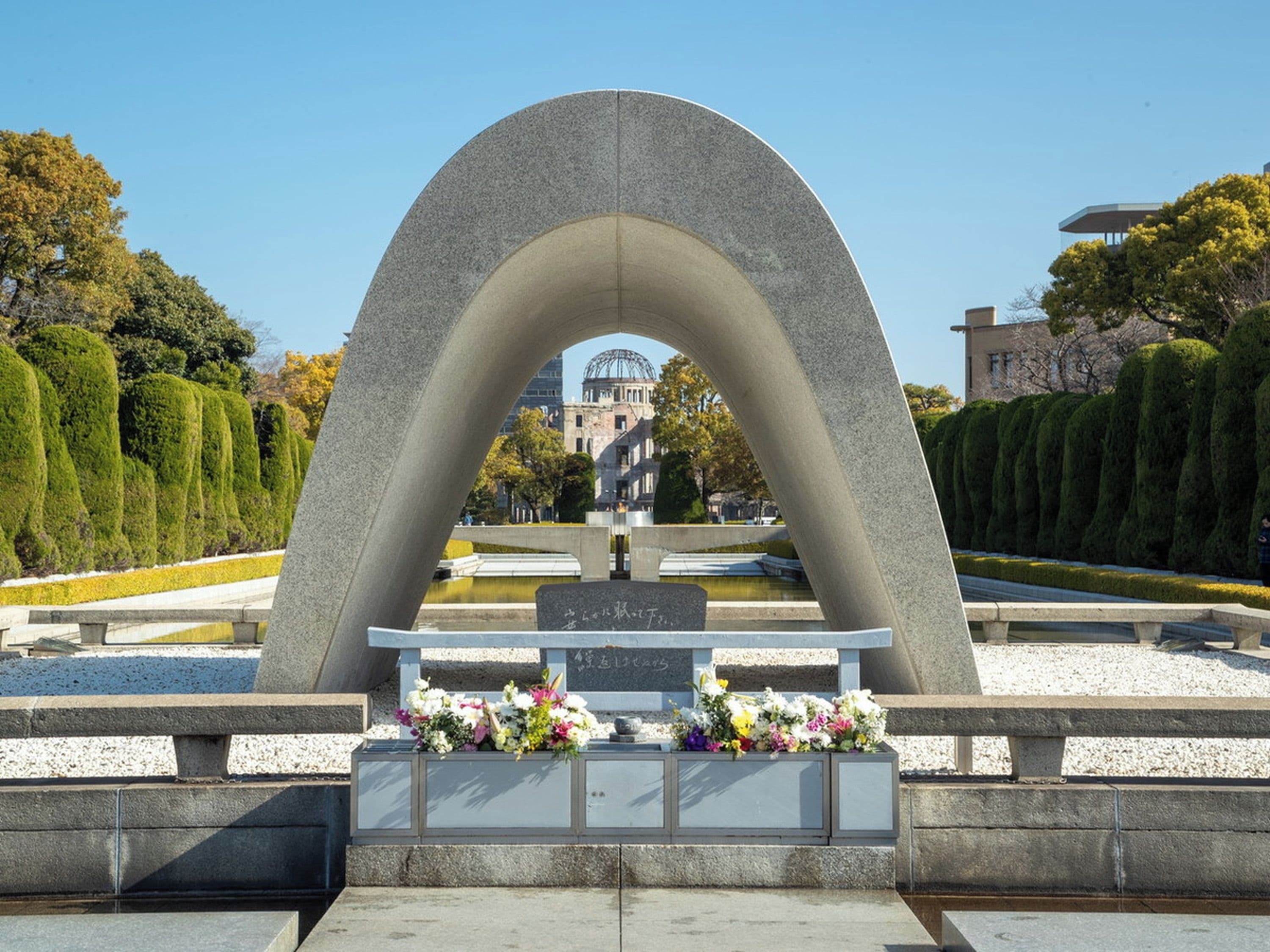 Monumento de pedra com inscrições e homenagens florais em um parque arborizado.