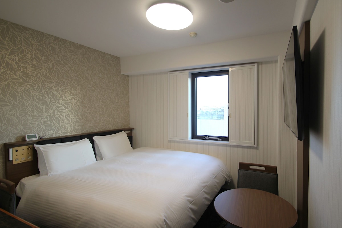 Una camera d'albergo con un letto, un comodino, una sedia, una finestra e una TV a schermo piatto fissata al muro.