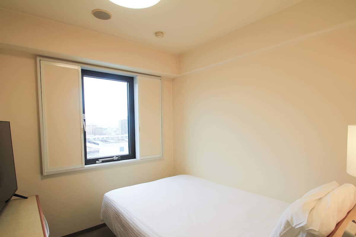A hotel room with a single bed, a window with blinds, a desk with a telephone, and a television.