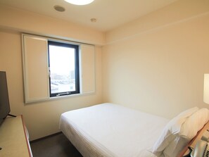 A hotel room with a single bed, a window with blinds, a desk with a telephone, and a television.