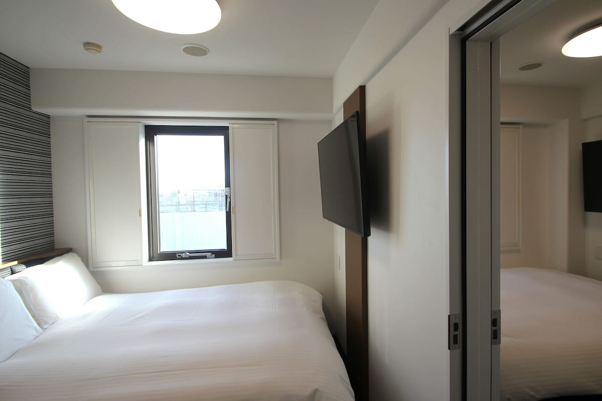 Una camera d'albergo con due letti, una TV a schermo piatto fissata al muro, una finestra con persiane e una lampada a soffitto.