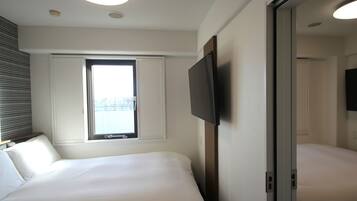 A hotel room with two beds, a flat-screen TV mounted on the wall, a window with blinds, and a ceiling light.