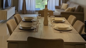 A dining area with a long table set for four, surrounded by chairs. There is a vase on the table, and the room has a modern design with recessed ceiling lights.
