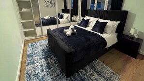 A bedroom with a black bed, white pillows, a blue rug, a wooden floor, a nightstand with a lamp, and a closet with shelves. - Luxury 5-bed Bungalow w/ Hot Tub, Games & Parking (Dudley)