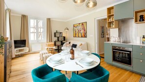 A modern living room with a round dining table, teal chairs, and a kitchen area with a built-in oven.