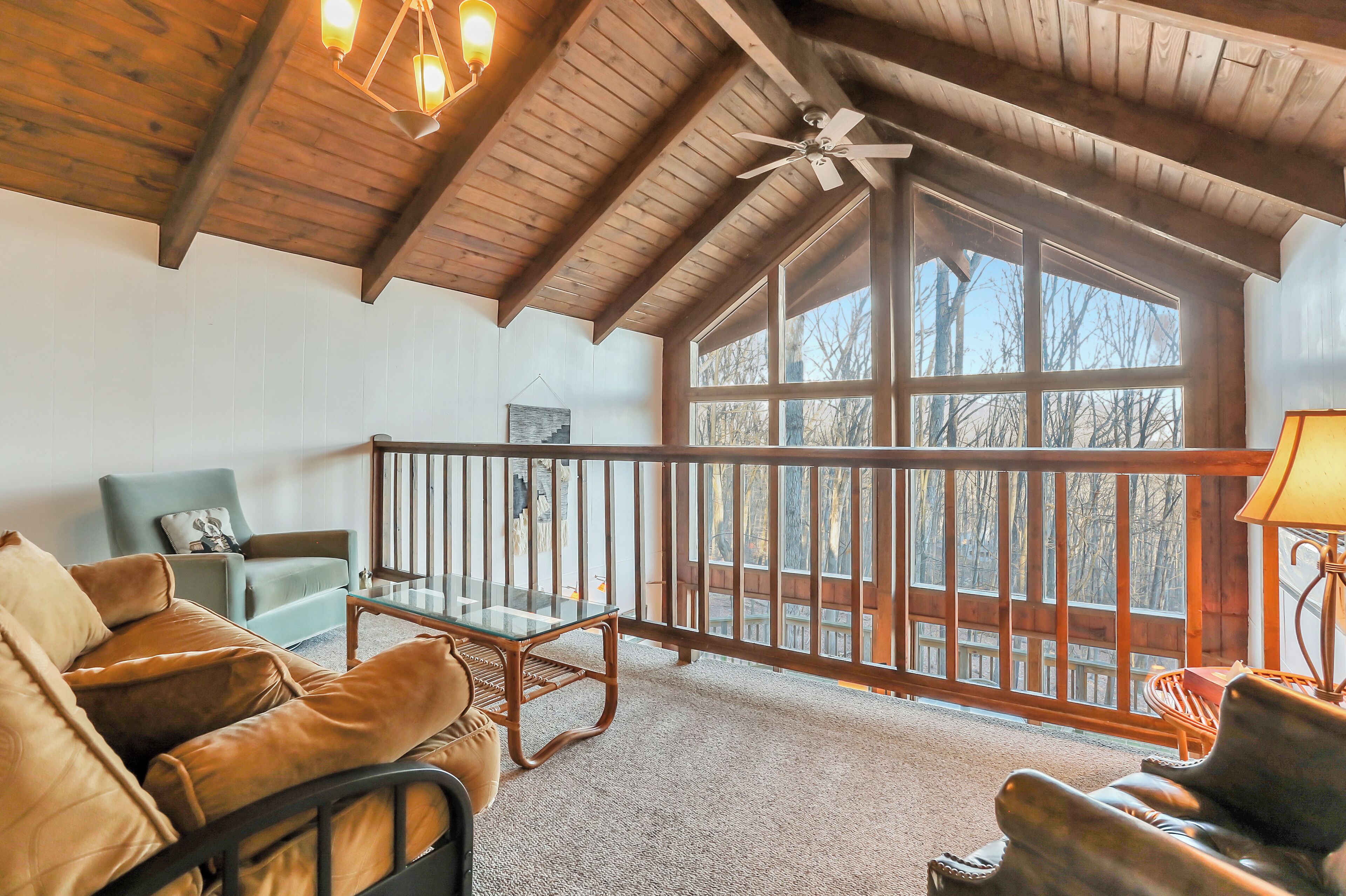 A cozy room with a wooden ceiling, a glass table, leather chairs, and a view of trees through large windows.