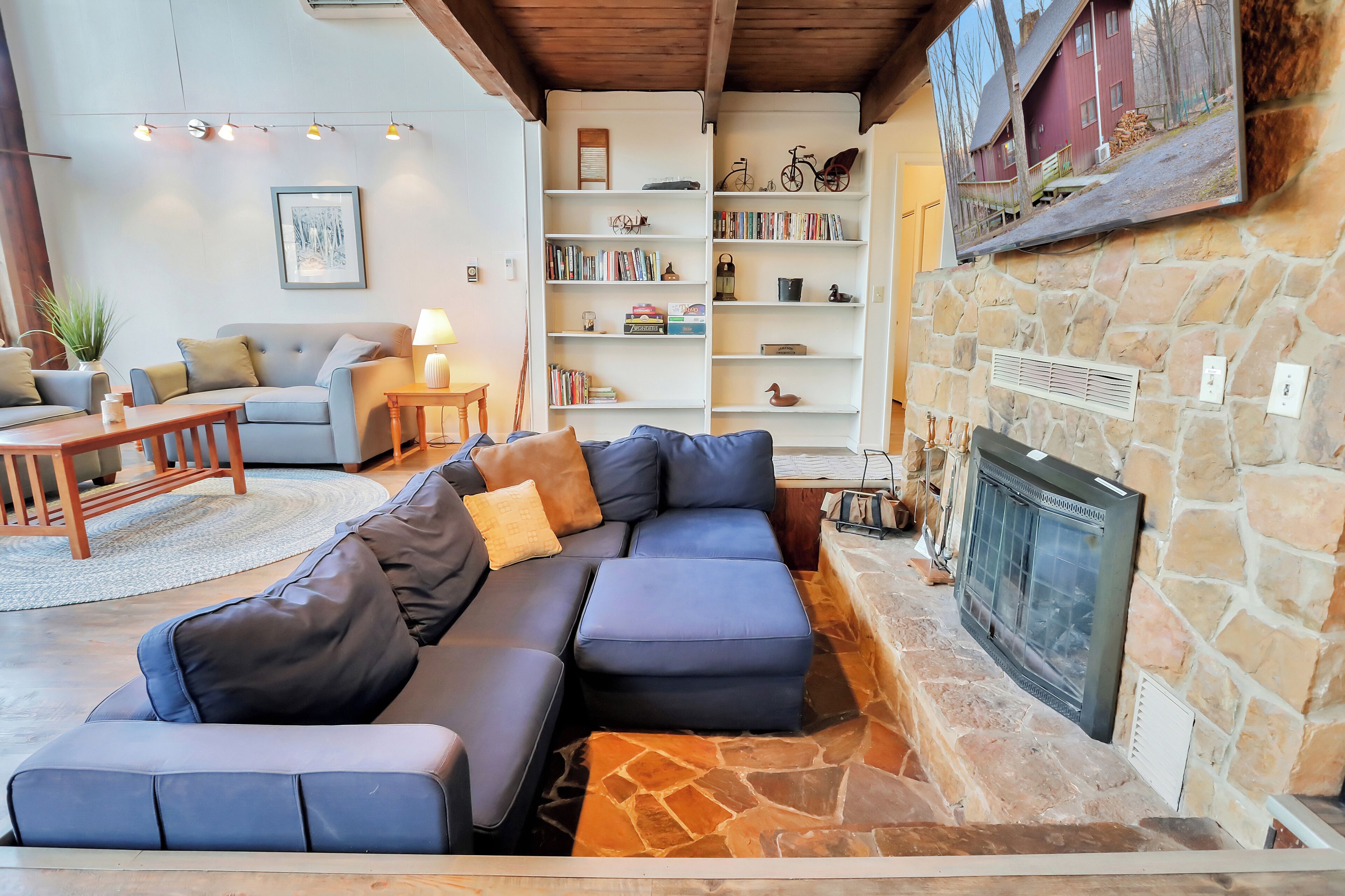 A cozy living room with a stone fireplace, a sectional sofa, a bookshelf, and a mounted television.