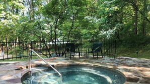 Un bain à remous en pierre, entouré d’une clôture métallique, installé dans un espace boisé, avec des chaises et une table à proximité.