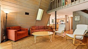 Una sala de estar acogedora con un sofá, un sillón y una mesita de centro, paredes de madera y una cocina al fondo.
