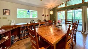 A spacious dining area with a large wooden table and chairs, surrounded by bookshelves and a view of the outdoors. - Wildflower- Themed | Pet Friendly | Private Hottub (Berkeley Springs)