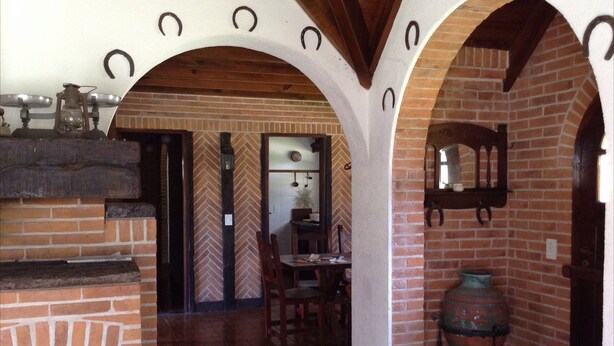 A rustic interior with brick walls, wooden beams, and a fireplace. There is a dining area with a table and chairs, and a large decorative pot on the floor.