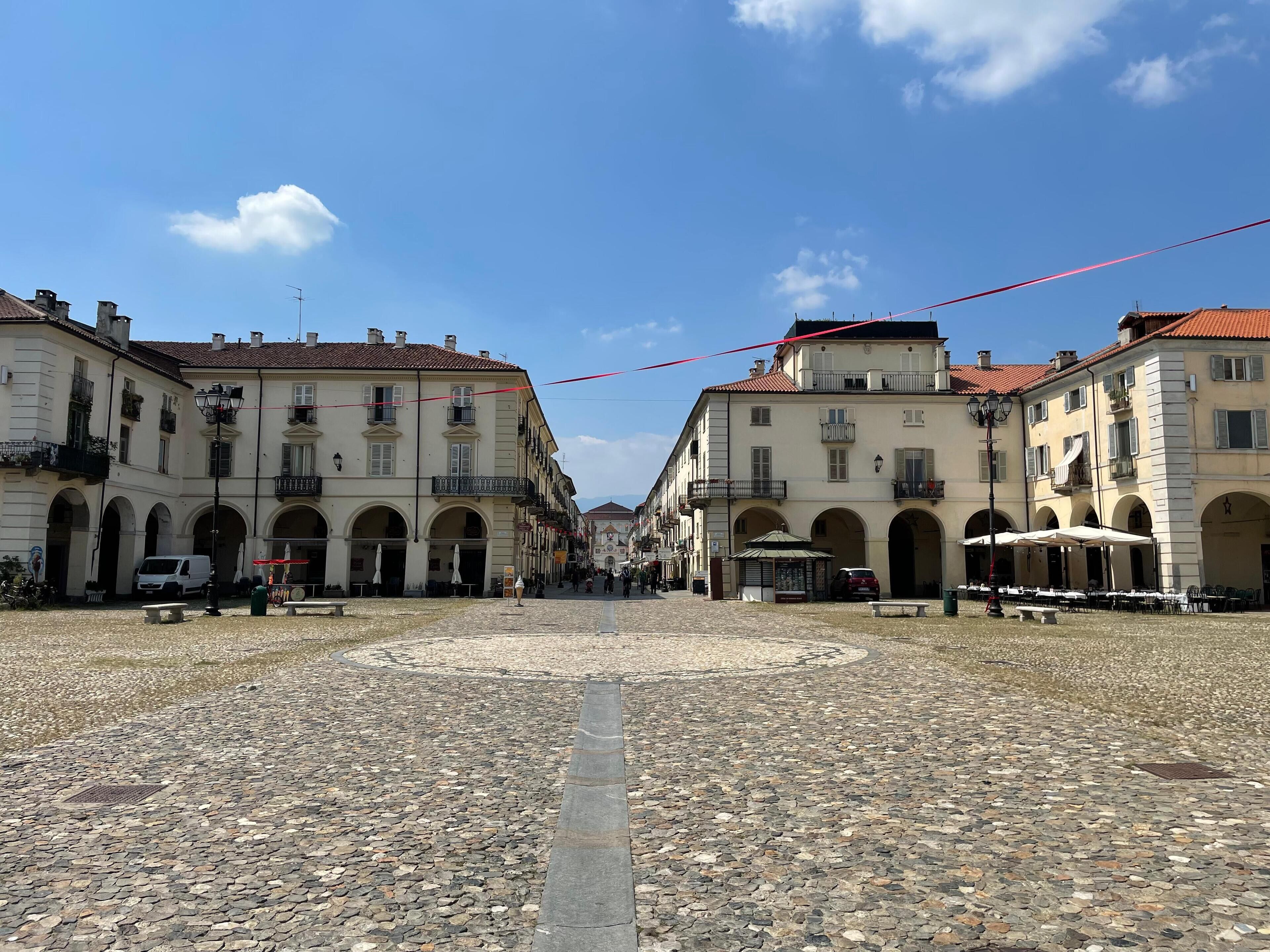 Ein Kopfsteinpflasterplatz mit historischen Gebäuden, Bogenpassagen und Sitzgelegenheiten im Freien.